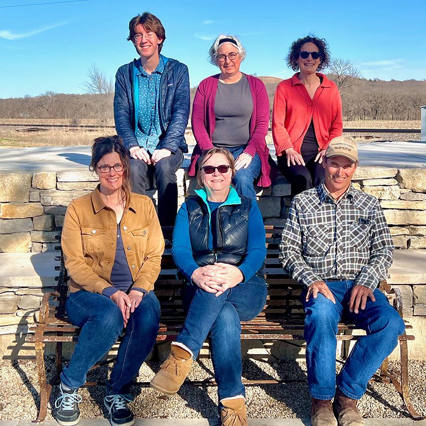 Group of six people smiling, sitting and standing outside on a stone wall on a sunny day.
