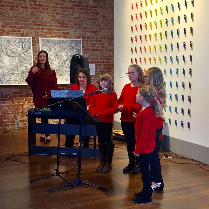 A group of children and adults sing in front of art and a colorful wall display.