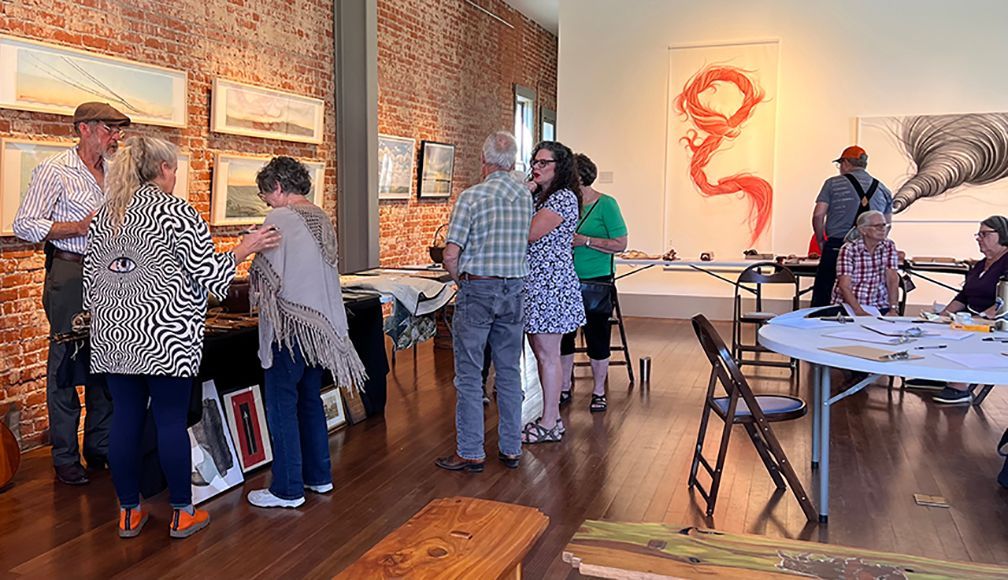People view art in a gallery: paintings on brick wall, large abstract art, tables, and wooden floors.