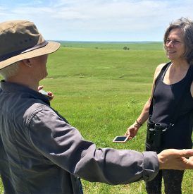 Two women in a grassy field, one gesturing, the other holding binoculars and a phone, on a sunny day.