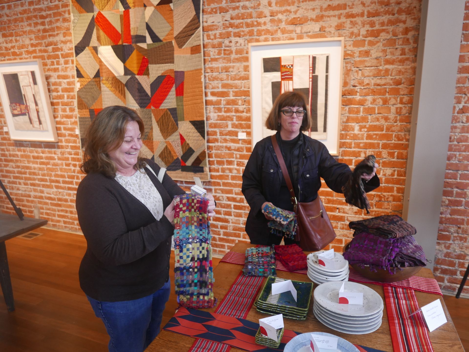 Two women at a craft fair display. Red brick wall background. One woman holding items, the other handling a scarf.