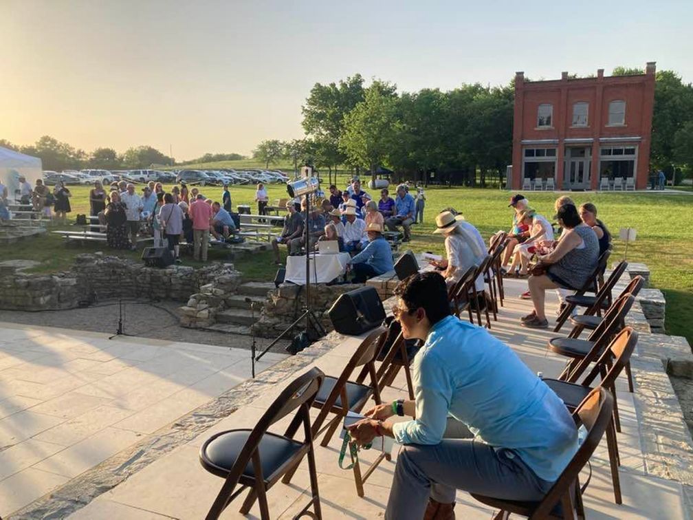 Outdoor event with people seated in chairs, stage with microphones, brick building in background.
