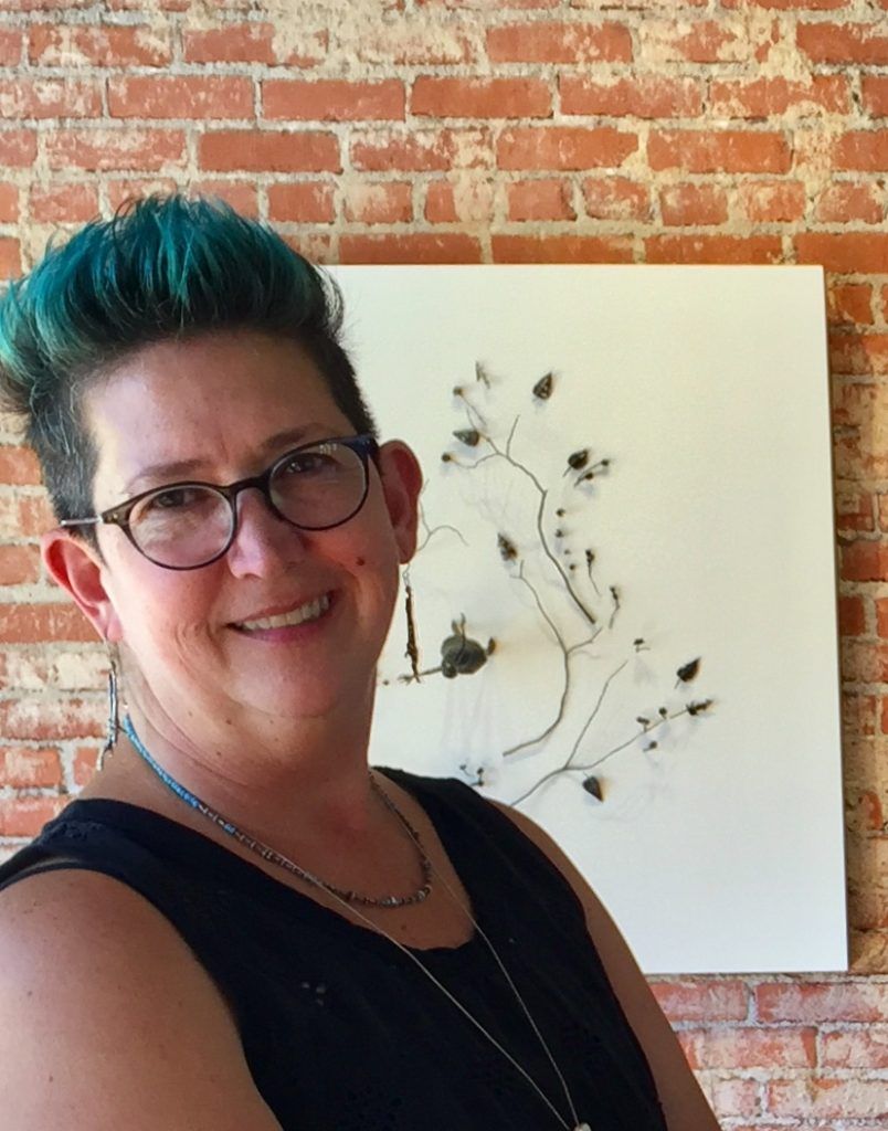 Woman with blue hair and glasses smiles in front of a brick wall and a white canvas with abstract art.