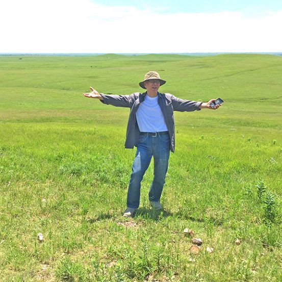 Man in field, arms outstretched, wearing hat, jeans, and jacket; green grass, blue sky.