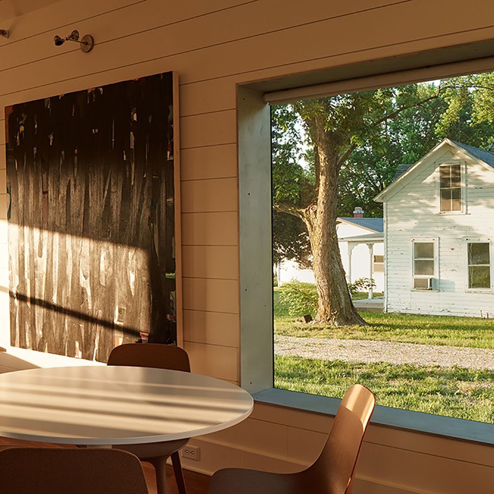 Interior view with dining table, artwork, and window overlooking a white house and tree.