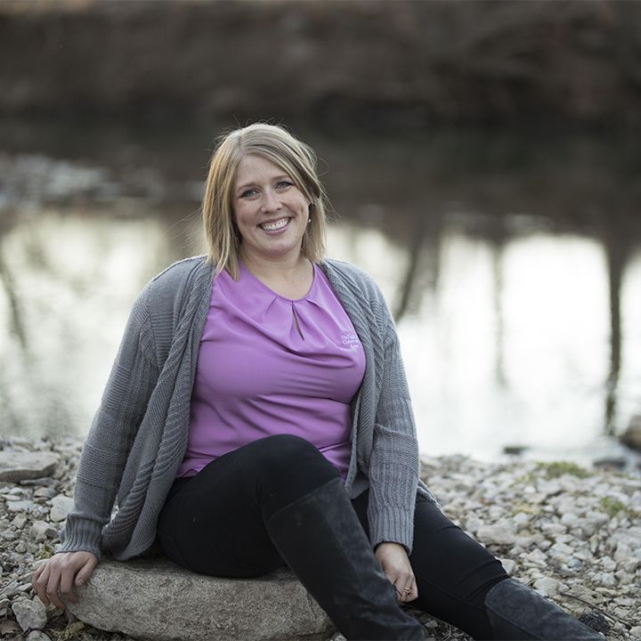 A woman in a purple shirt and gray sweater is sitting on a rock.