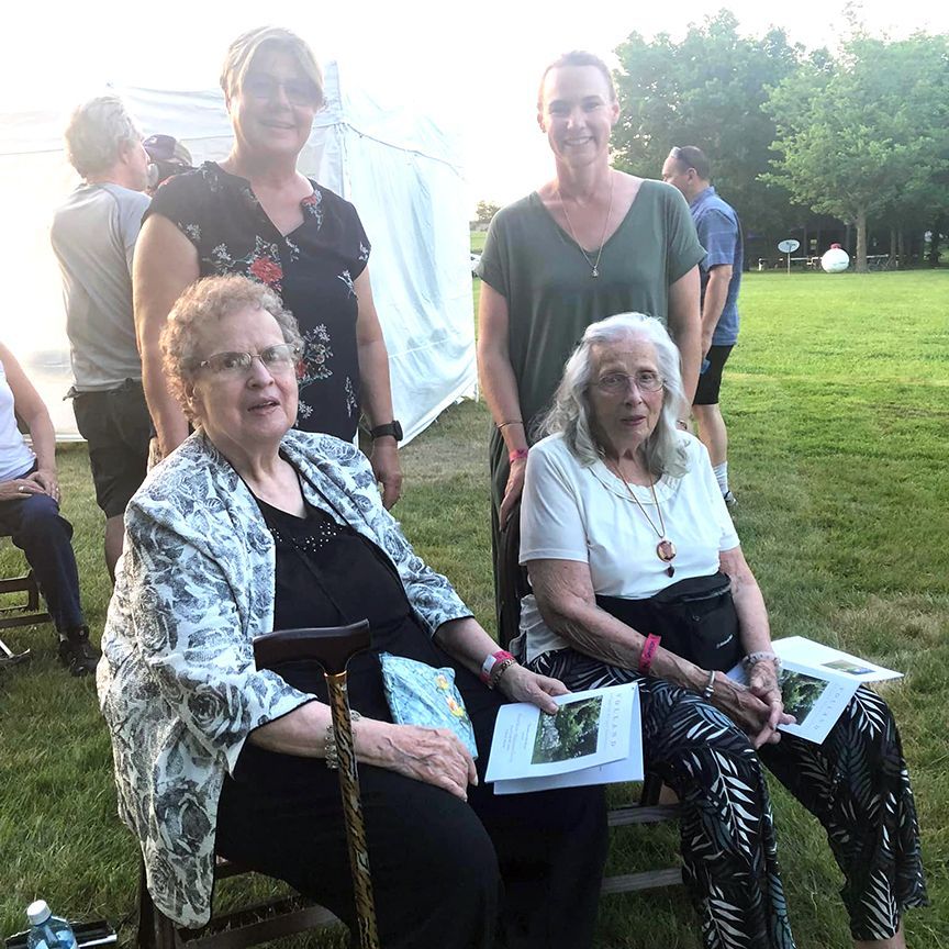 Group of five people at an outdoor event, two elderly women seated, holding books, with three women standing behind them.