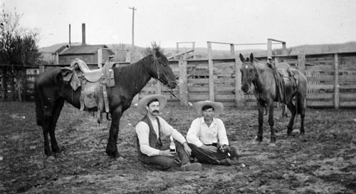 Two men are sitting on the ground next to horses in a black and white photo.