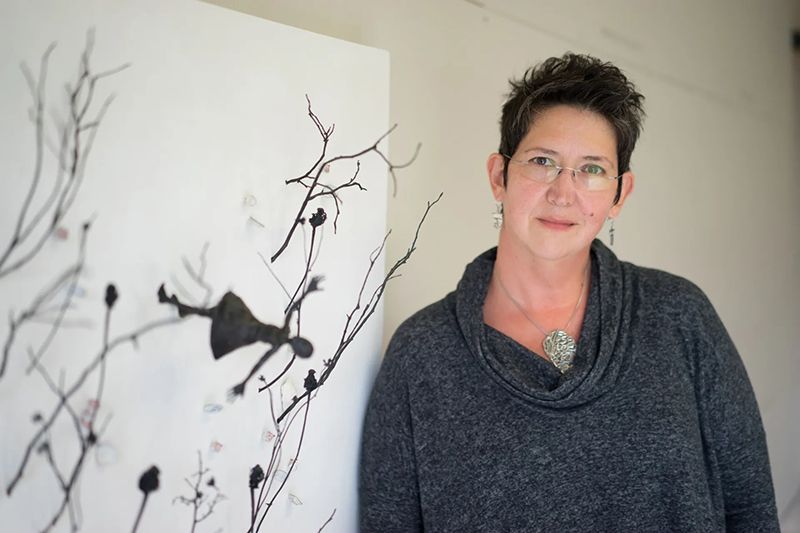 Woman with short dark hair, glasses, and grey sweater poses next to a black and white art piece of plant branches.