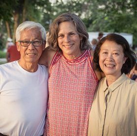 Three people smiling for a photo: an older Asian man and woman flanking a middle-aged woman outdoors.