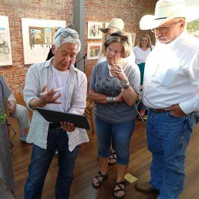 A group of people in an art gallery, looking at artwork. Man in cowboy hat, woman in t-shirt and jeans, and another man holding a tablet.