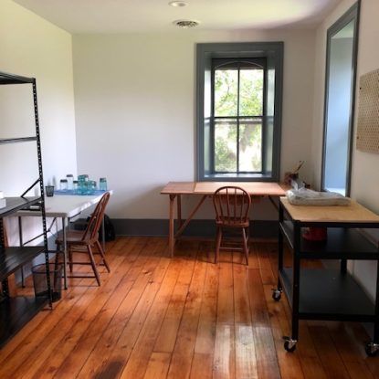 Room with wooden floor, white walls, and a window. Work tables and chairs are visible.