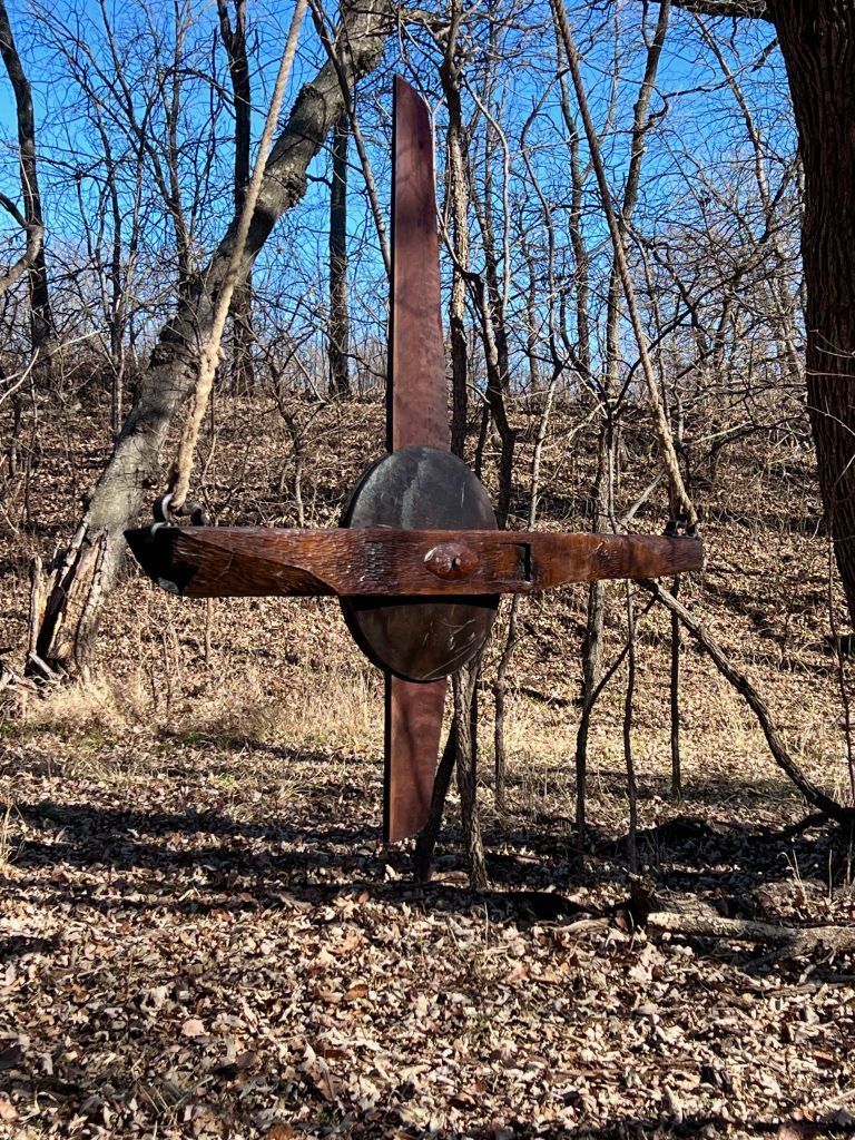 Rusty metal sculpture resembling a cross in a wooded area, brown with a blue sky background.