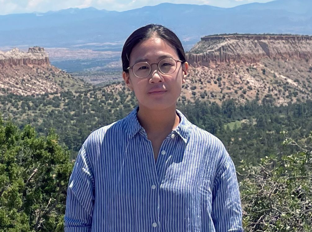 Woman with glasses in blue striped shirt, scenic desert landscape background.
