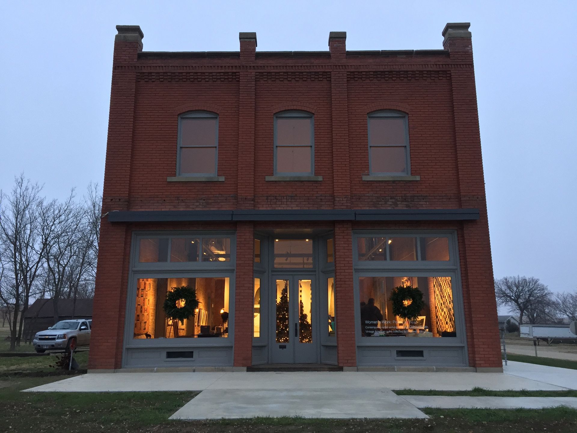 Brick building with large windows, lit interior, wreaths, and cloudy sky.