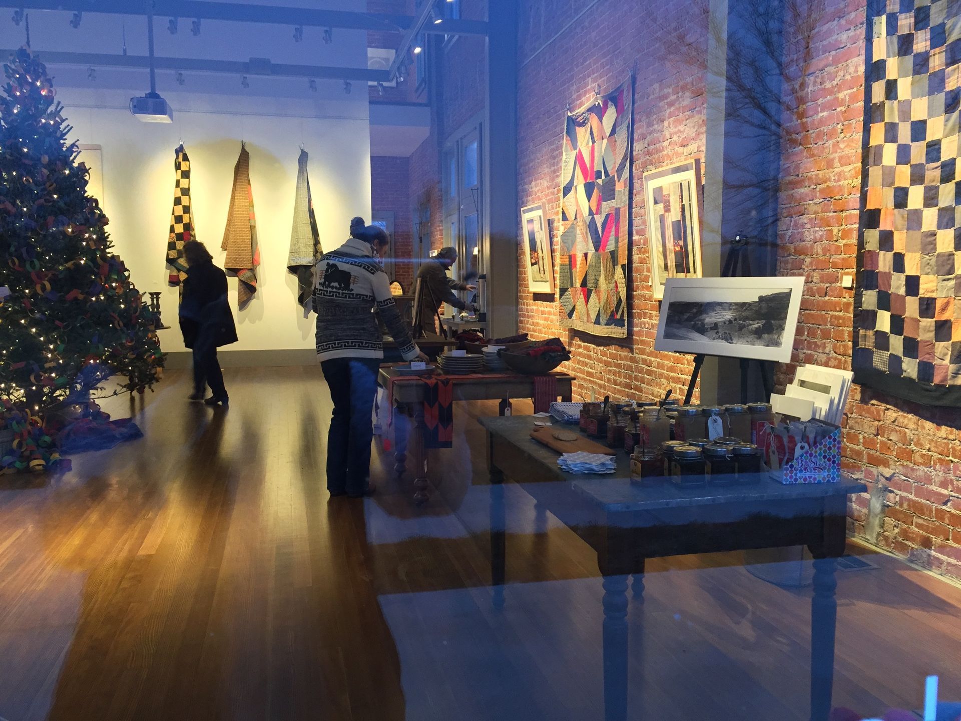 Inside an art gallery, people browse art near a Christmas tree. Wood floors, brick walls, and quilts.