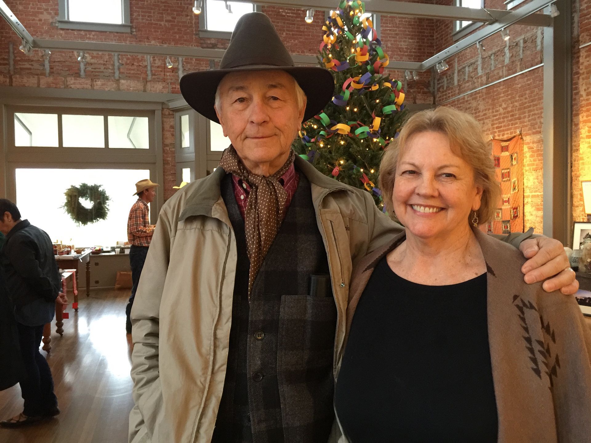 Man in hat and woman smiling, arm around each other near a Christmas tree in a brick-walled room.