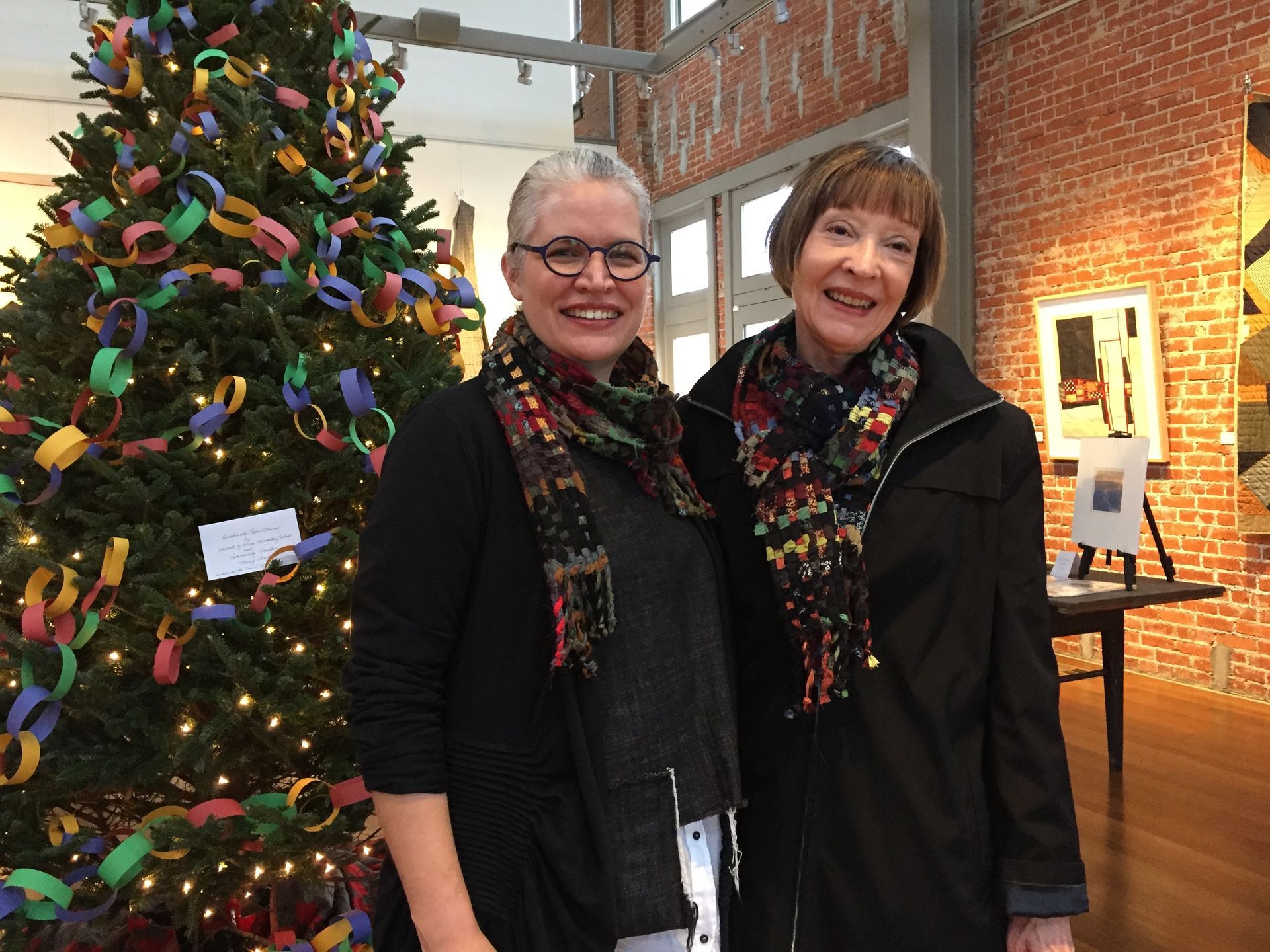 Two women smiling in front of a decorated Christmas tree in an art gallery.