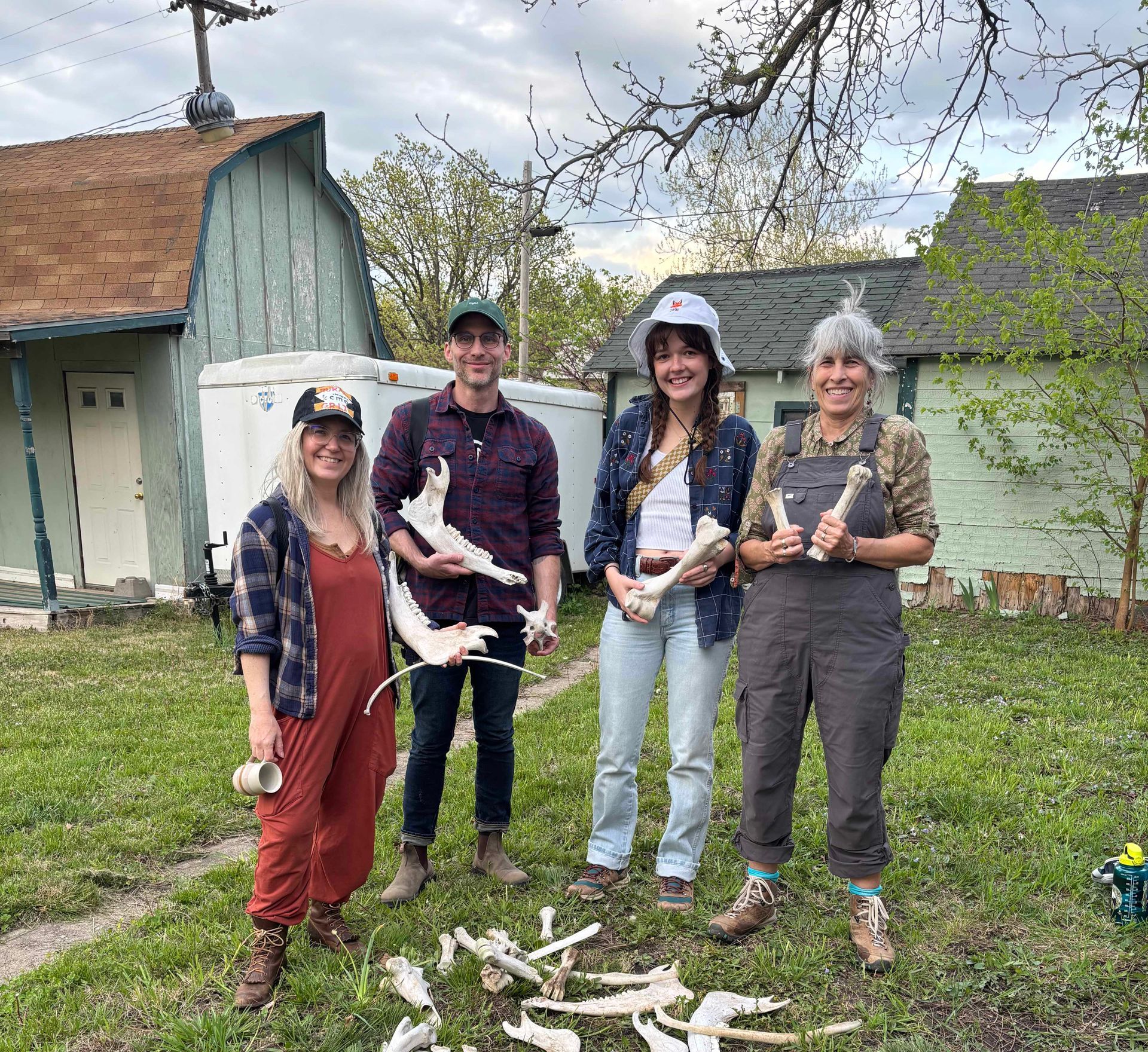 Four people holding animal bones outside. They stand in front of houses with green and brown exteriors, grass and scattered bones on the ground.