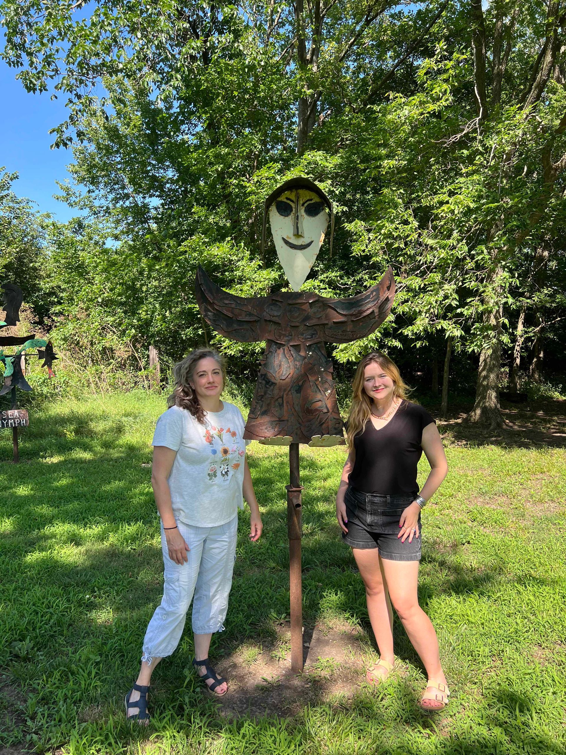 Two women pose with a metal sculpture in a green, sunny outdoor setting.
