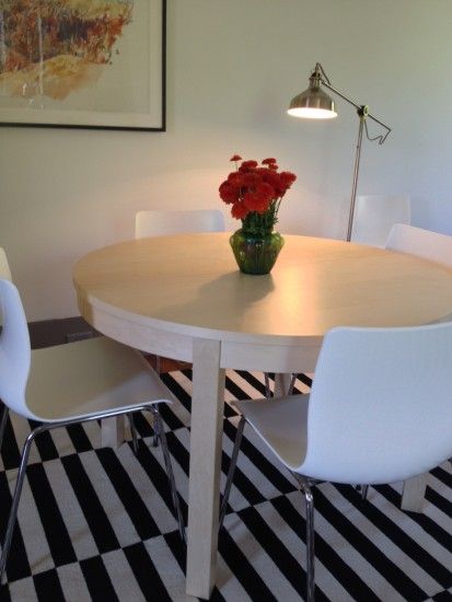 Round wooden table with red flowers, white chairs, black and white striped rug, lamp.