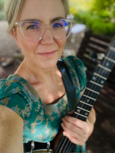 Woman with glasses holding a banjo outdoors; green dress, blurred background.