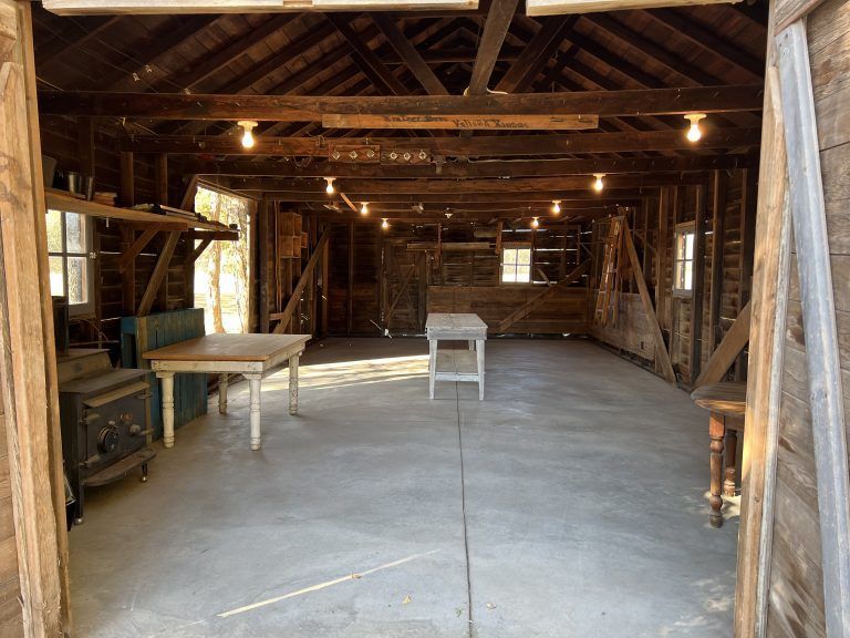 An open wooden garage with concrete floor. Two tables and a wood-burning stove are inside, lit by hanging bulbs.