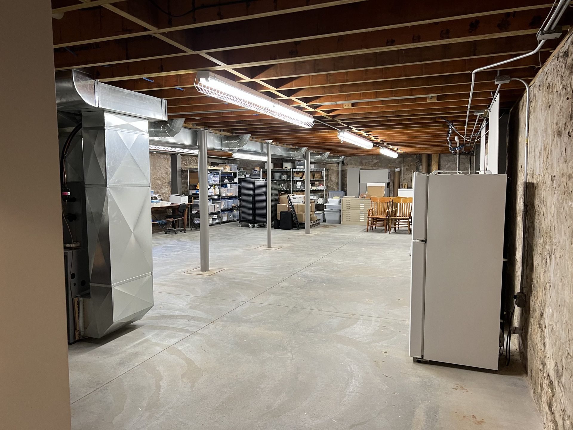 Basement with concrete floor, exposed ceiling beams, ductwork, refrigerator, and shelving.