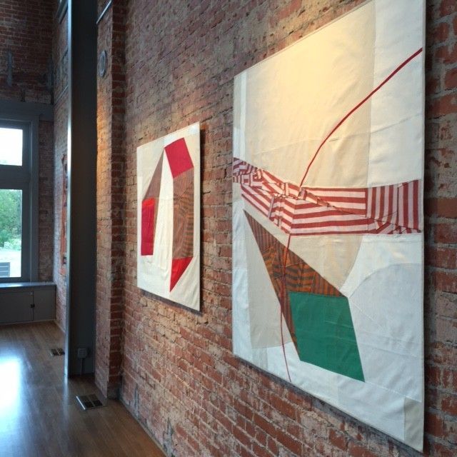 Two abstract fabric artworks hang on a red brick wall in a gallery.
