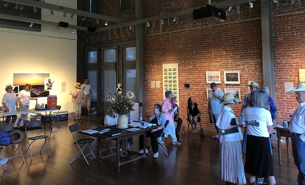 People gather in a gallery with brick walls, viewing artwork. Sunlight streams in from windows.