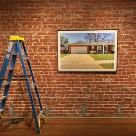 Blue ladder leans against a red brick wall. A framed photo of a house hangs to the right.