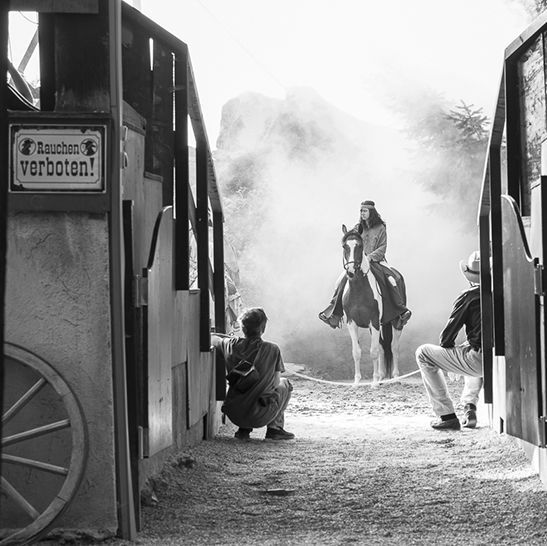 Black and white scene: Person on horseback amidst smoke, between wooden structures, two figures watching.