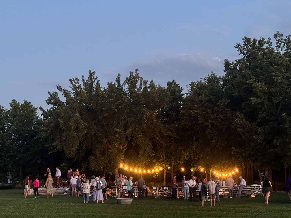 a group of people gather under warm lights outside. there is an open theater and sunset light in the background.