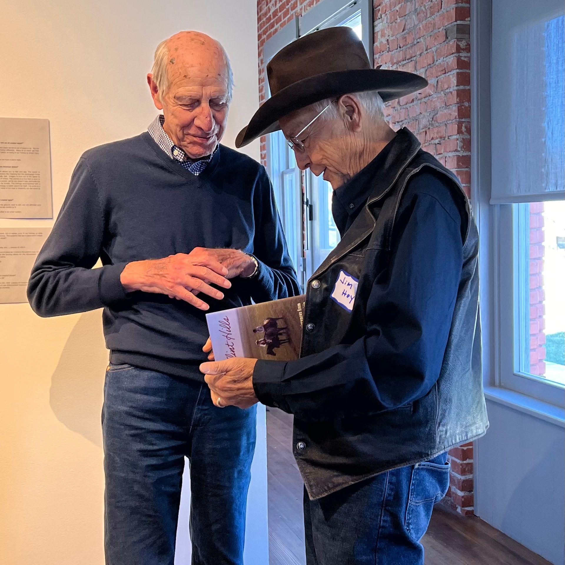 Two older men examining a book in a gallery. One wears a hat and vest. Natural light.