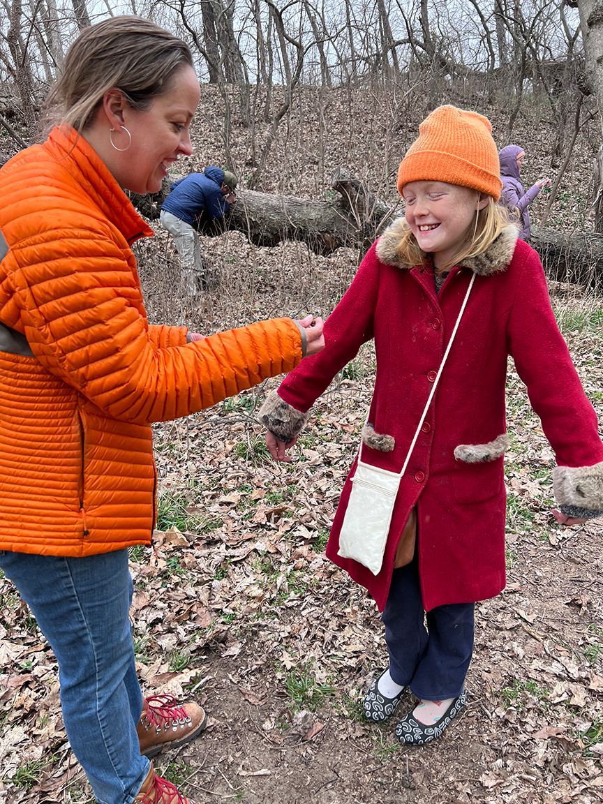 Woman in orange jacket holding hands with smiling child in red coat and orange hat, outdoors.