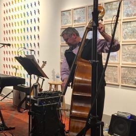 Man playing a double bass in an art gallery, near a keyboard and colorful wall art.