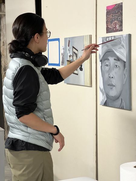 Woman painting a portrait on a wall, wearing a vest and glasses. Other paintings visible nearby.