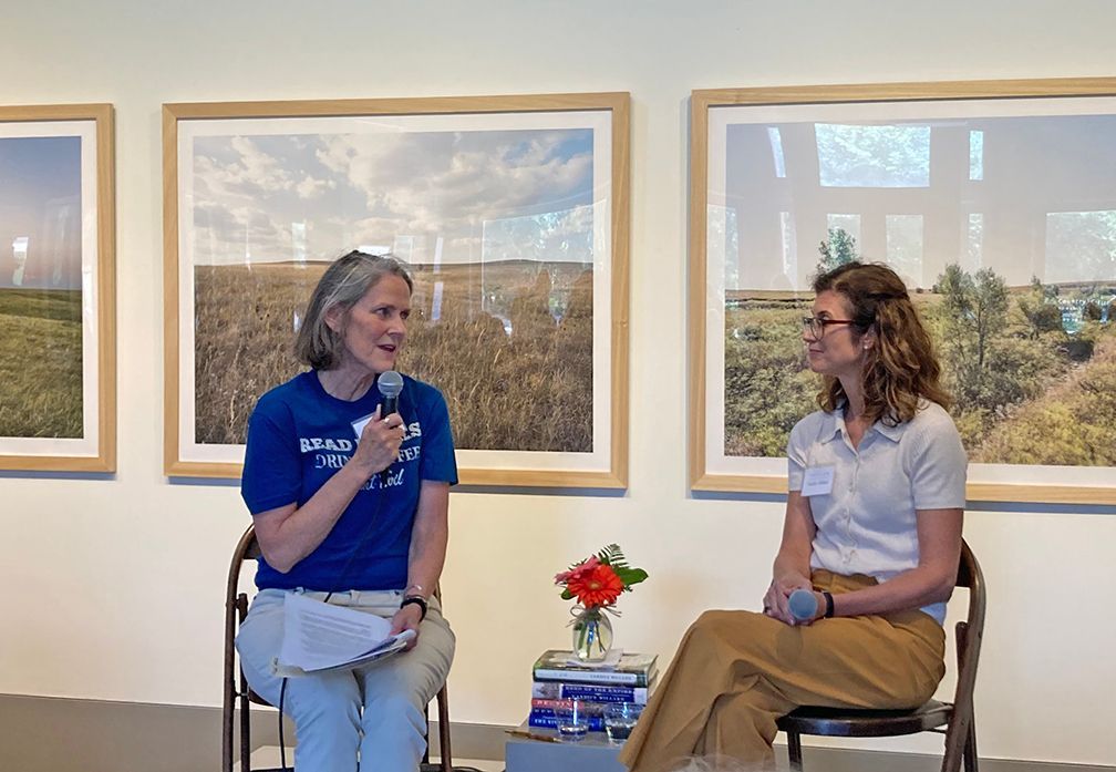 Two women in a gallery, one speaking into a microphone. They sit with a small vase of flowers and books, framed photos behind them.