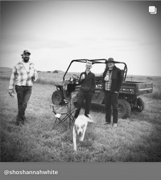 Three people and a dog by a utility vehicle on a field. One person smiles, holding a drink. Overcast sky.