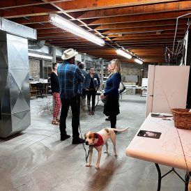 People in an art studio, with a dog on a leash. Exposed ceiling beams and ductwork.