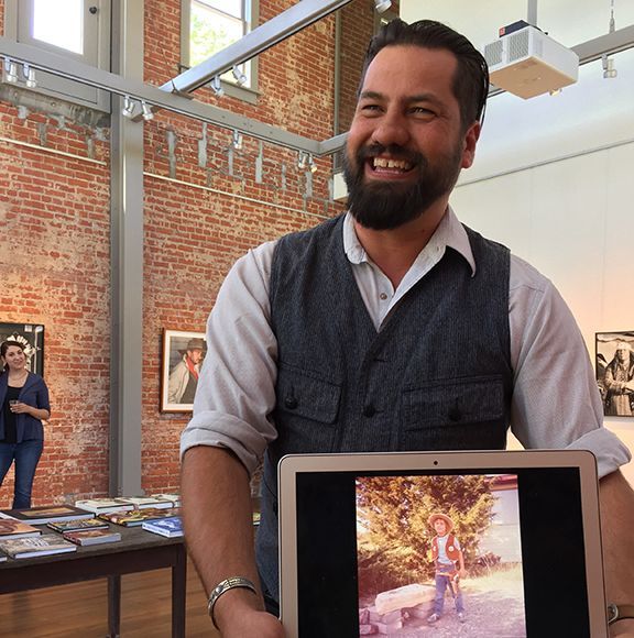 Man smiles, holding laptop showing childhood photo. Art gallery: brick wall, tables of books, other people present.