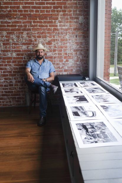 Man in cowboy hat sits by window with photos on display. Brick wall background.