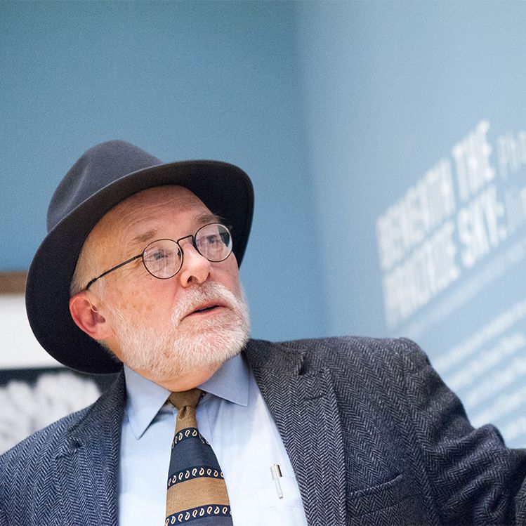 Man with glasses and hat, looking up and right, in front of a blue wall with text.