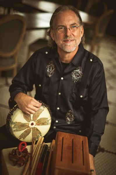 Man with glasses, smiling, holding a drum and percussion instruments. Sitting in a tiled room.