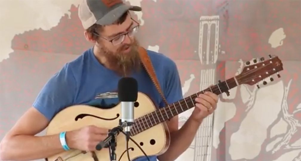 Man with a beard playing an eight-stringed guitar, wearing glasses and a hat, in front of a mural.