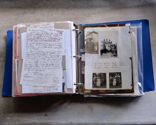 An open blue binder filled with old photos, handwritten notes, and newspaper clippings on a table.