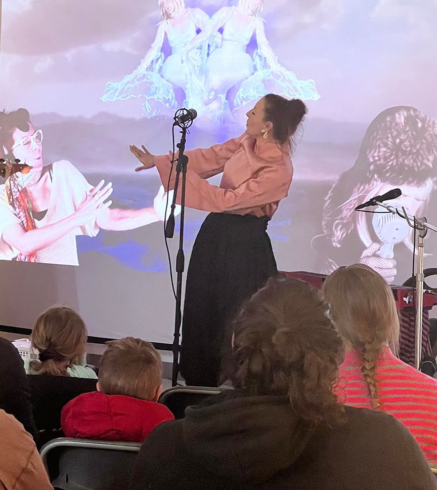 Woman performs on stage with projected imagery; audience watches.