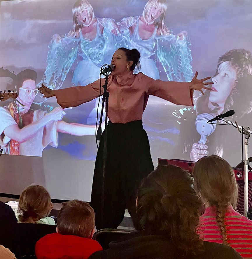A woman singing onstage with arms outstretched; audience in foreground, background features video with figures.