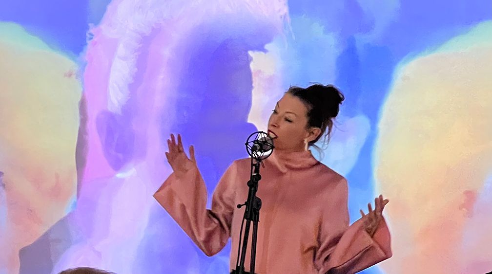 Woman singing into a microphone, arms raised, against a colorful abstract background.