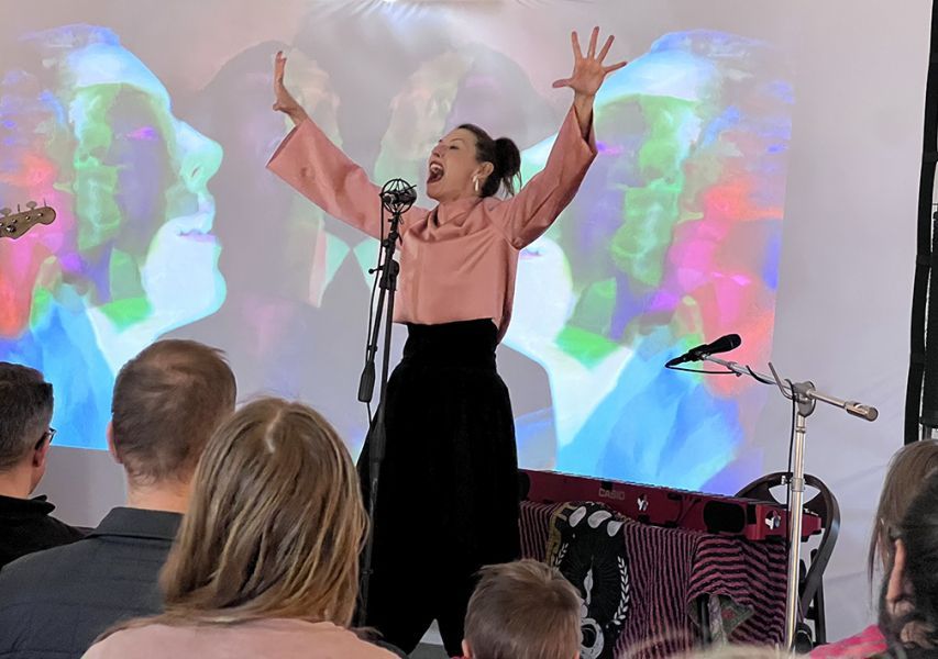 Woman singing with arms raised, performing in front of a colorful projected image. Audience seated in a venue.